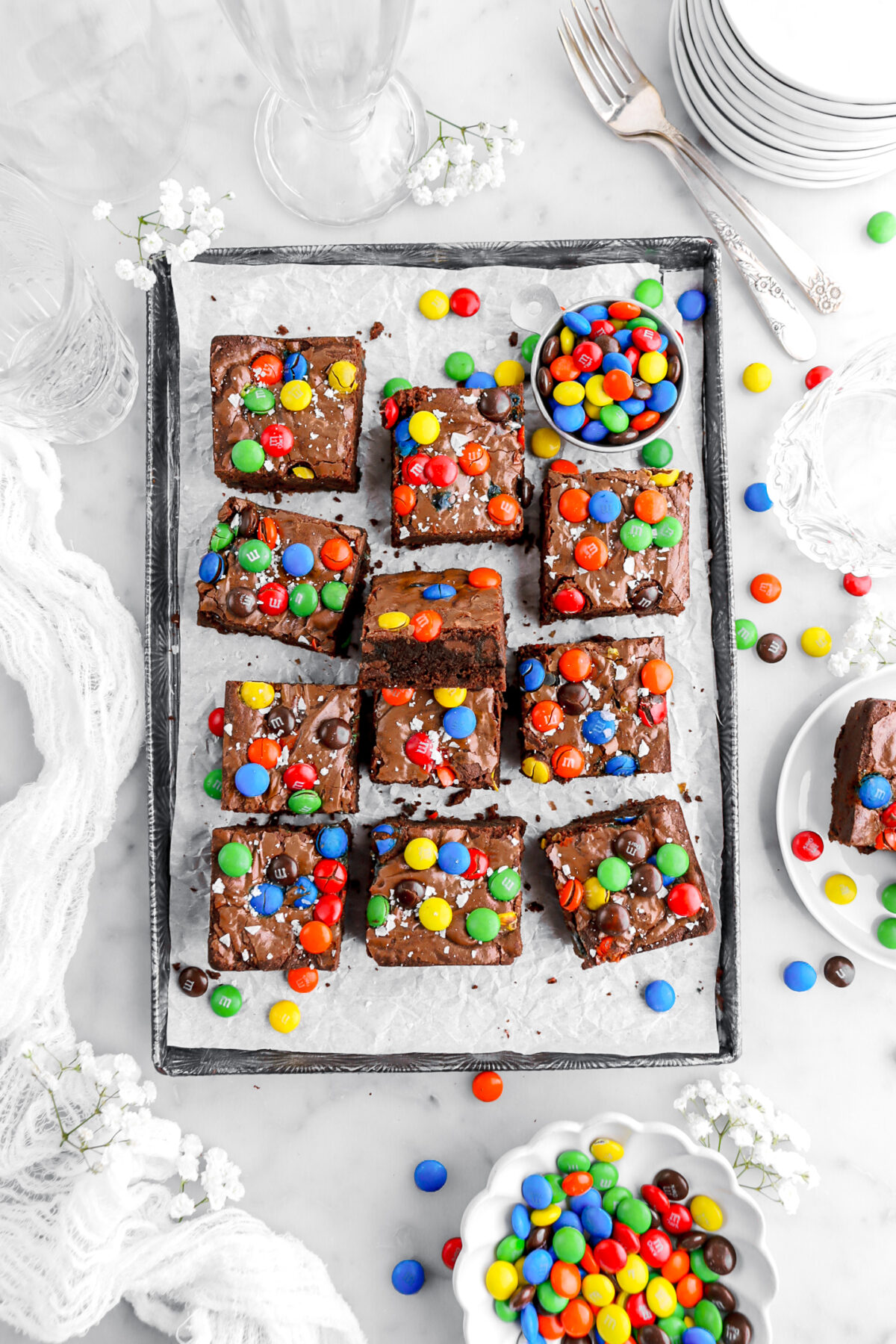 M&M brownies on lined sheet pan with two bowls of M&M's and more candies scattered around on marble surface.