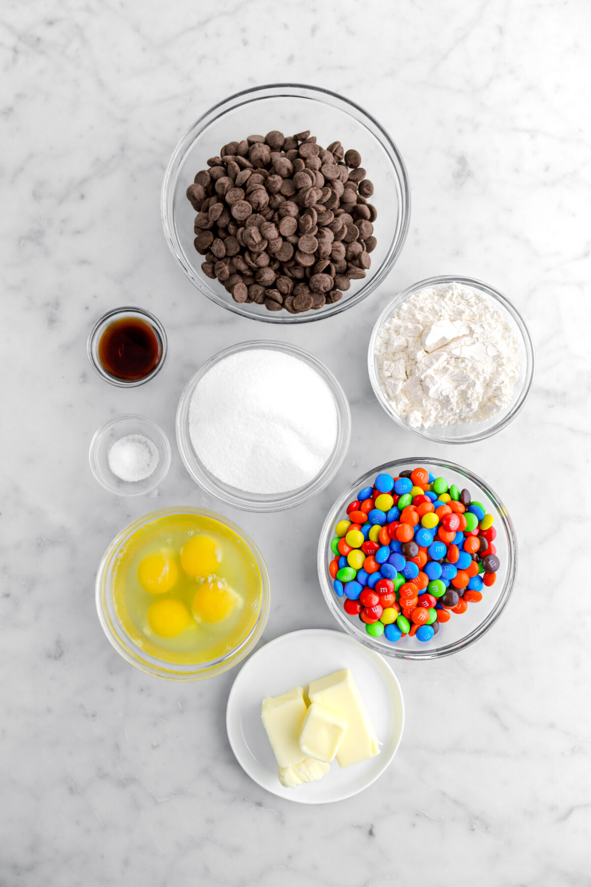 chocolate chips, vanilla, sugar, flour, M&M's, eggs, salt, and butter on marble surface.