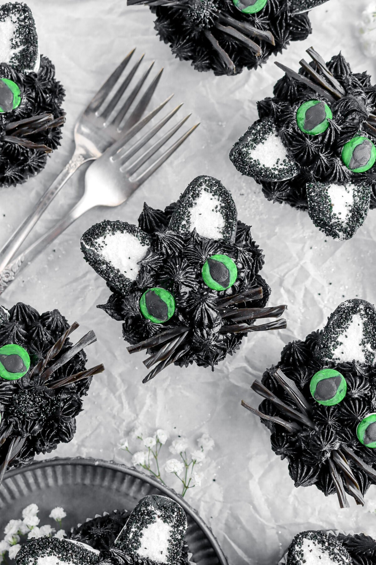 overhead image of black cat cupcakes on parchment paper with two forks and white flowers around.