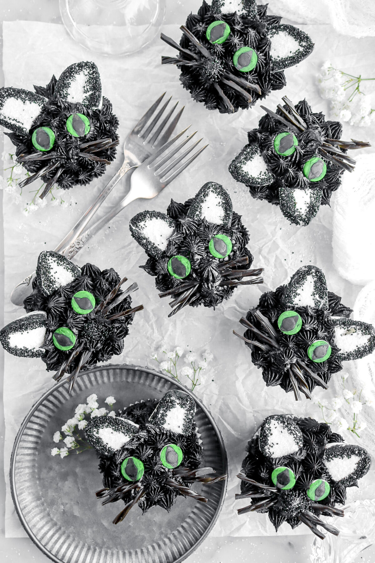 overhead image of eight black cat cupcakes on parchment paper with flowers, two forks around with one cupcake on metal plate.