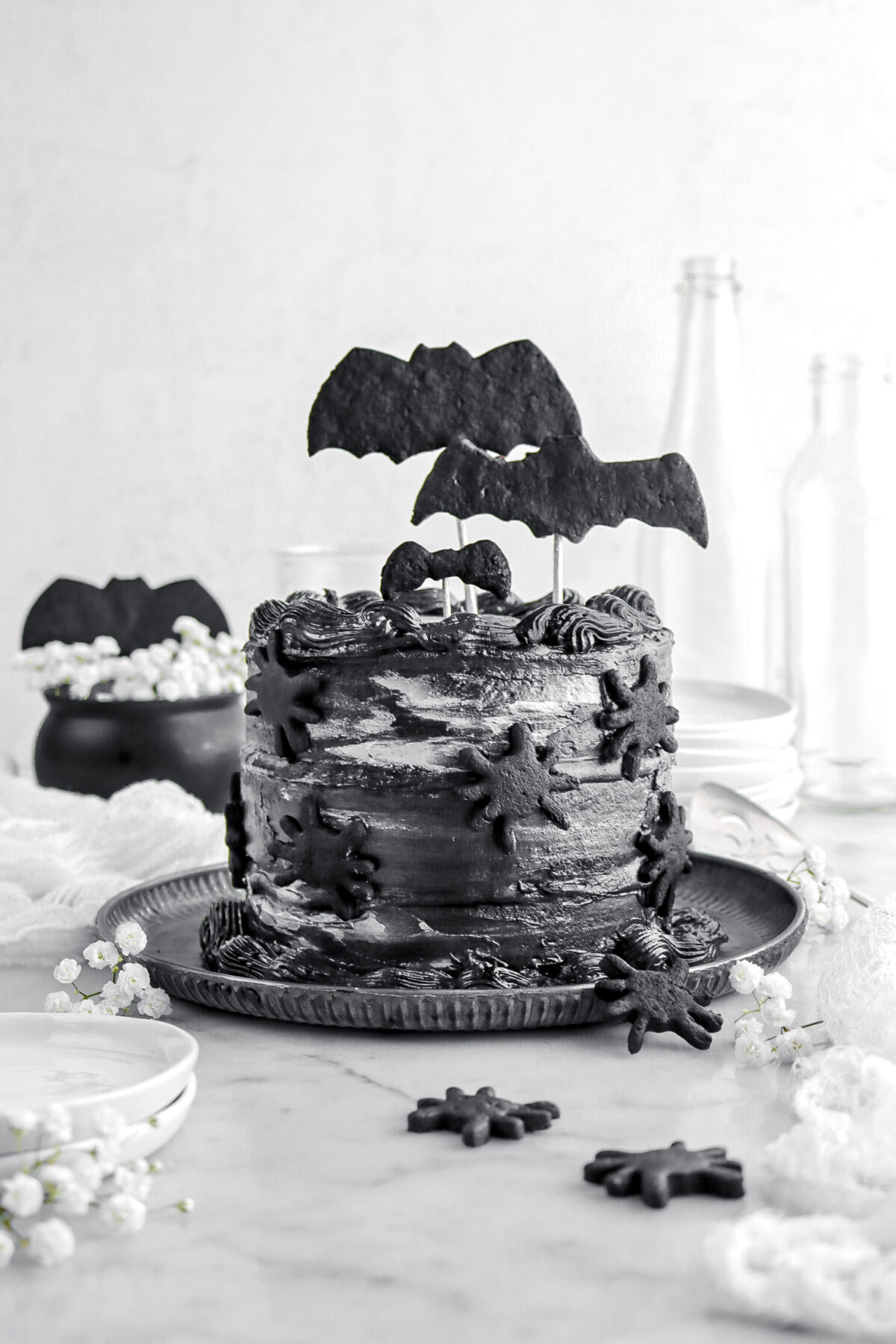 black cake with bats cookies on top and spiders on the side on metal plate with white flowers around and white cheese cloth, black cauldron behind, and empty glasses.