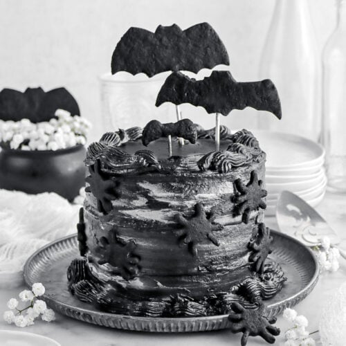 black velvet cake with black bat cookies on top and black spiders on the side with white flowers around, a stack of plates behind, and empty glasses on marble surface.