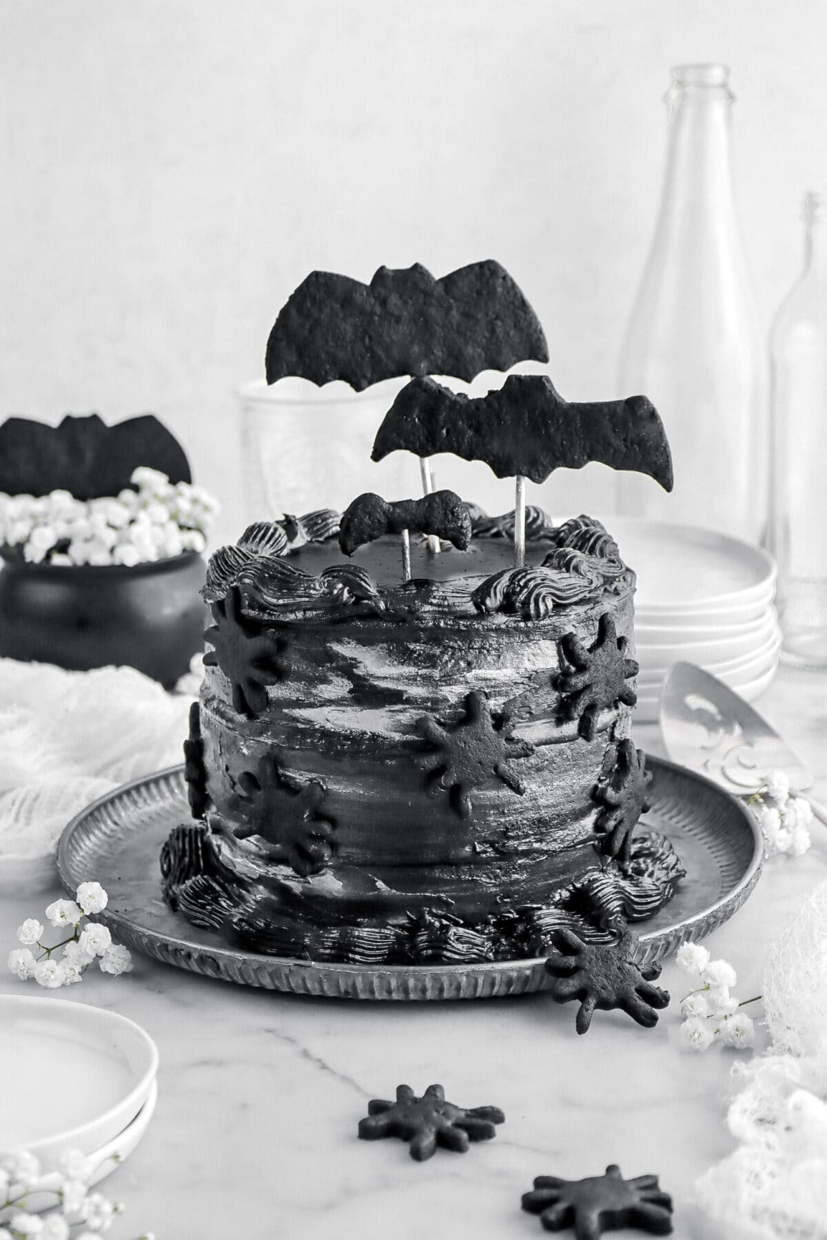 black velvet cake with black bat cookies on top and black spiders on the side with white flowers around, a stack of plates behind, and empty glasses on marble surface.