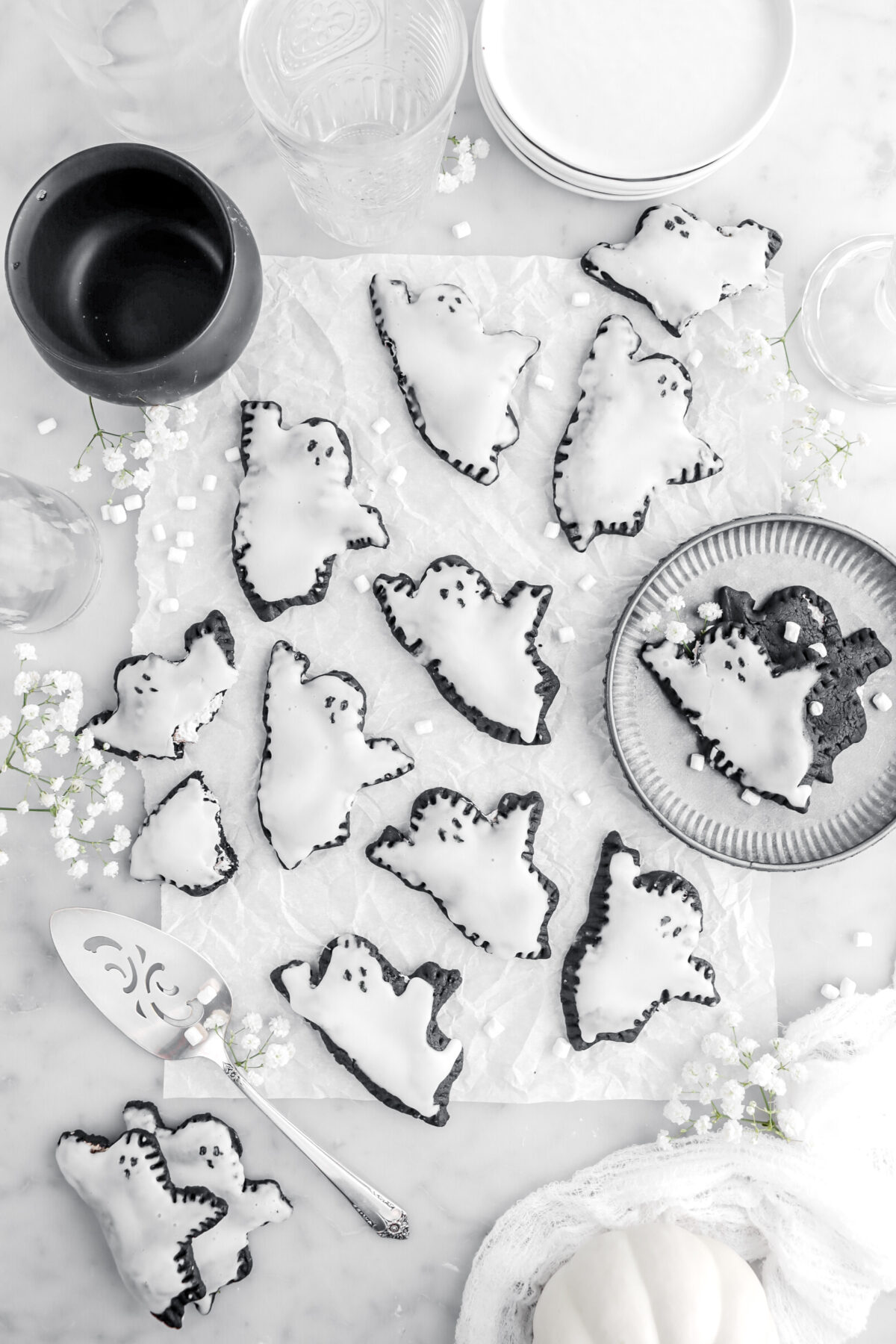 overhead image of ghost pop tarts on parchment paper with mini marshmallows and white flowers around, a black cauldron, stack of plates, and empty glasses on marble surface.