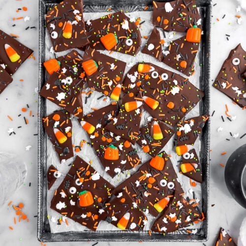 halloween candy bark on parchment lined sheet pan with extra sprinkles around and more pieces of candy bark on marble surface.