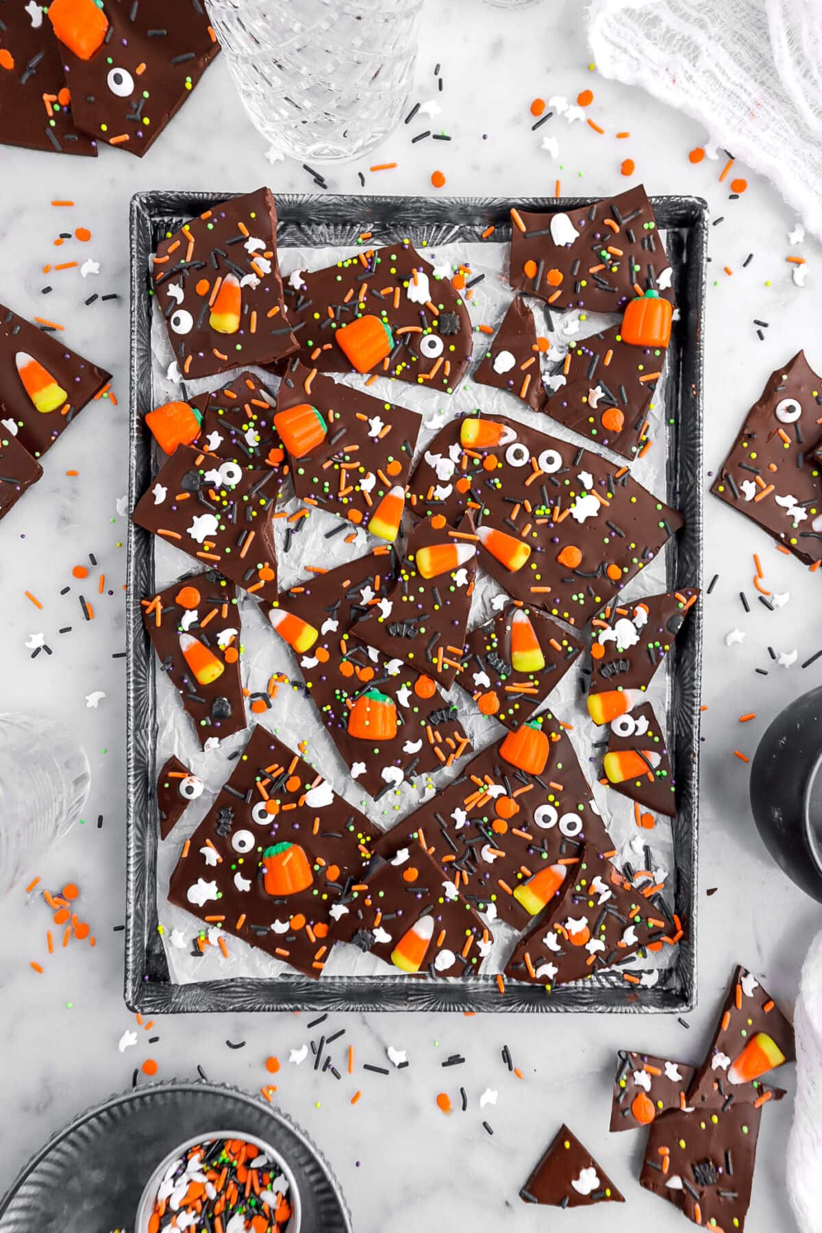 halloween candy bark on parchment lined sheet pan with extra sprinkles around and more pieces of candy bark on marble surface.