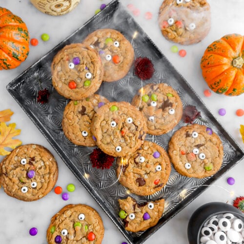 monster cookies on sheet pan with smoke and fairy lights, m&m candy, candy eyes, and pumpkins around on marble surface.
