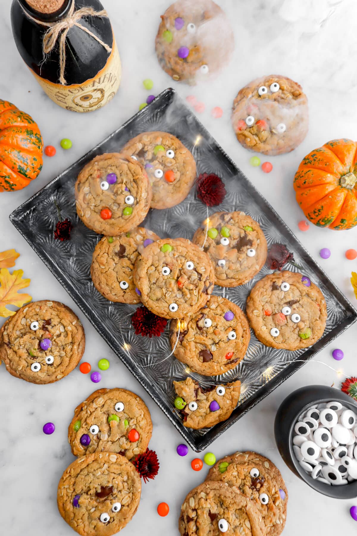 monster cookies on sheet pan with smoke and fairy lights, m&m candy, candy eyes, and pumpkins around on marble surface.