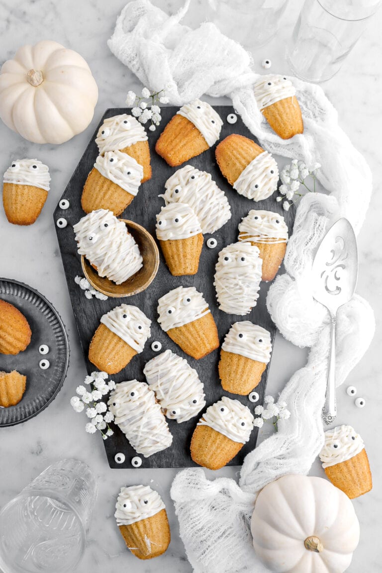 overhead image of mummy decorated madeleines on coffin shaped wood board with white flowers and candy eyes around, a white cheese cloth, and white pumpkins around on marble surface.