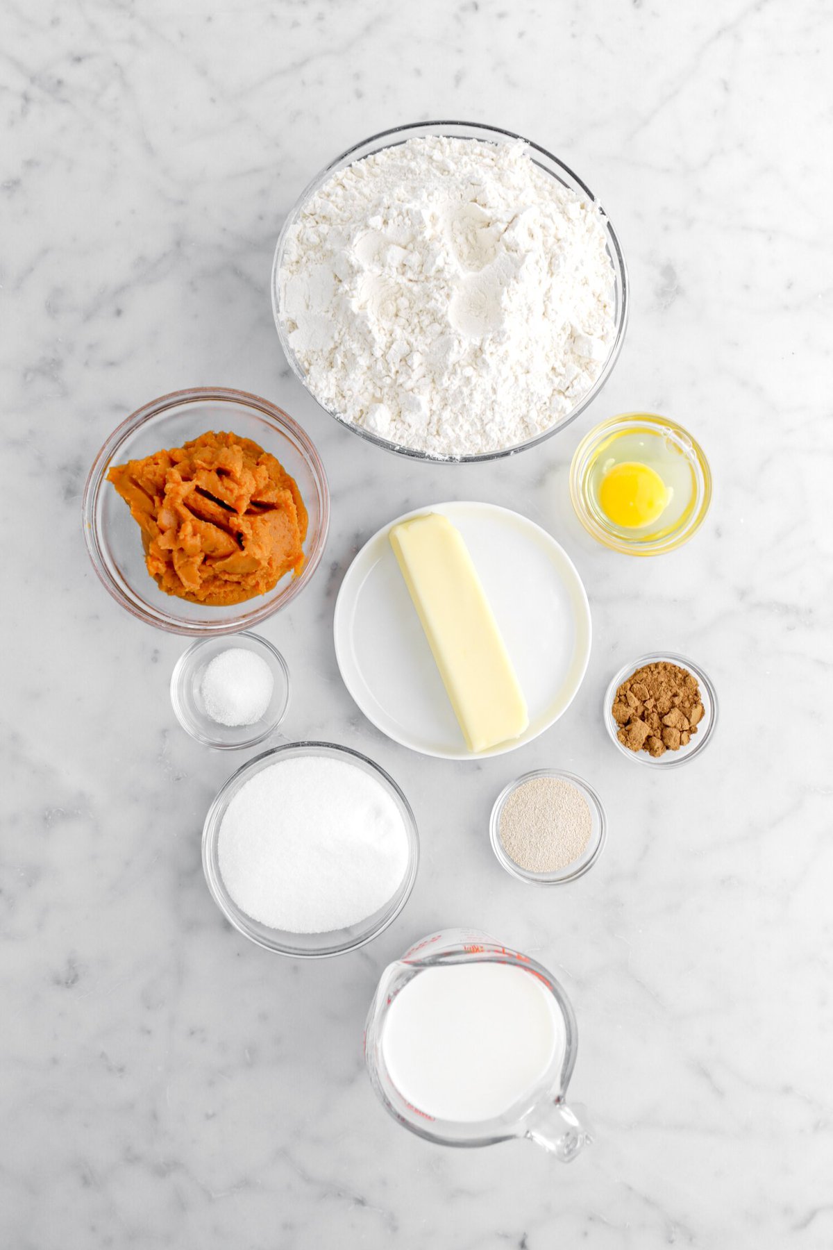flour, pumpkin puree, egg, stick of butter, salt, sugar, yeast, spice, and milk on marble surface.