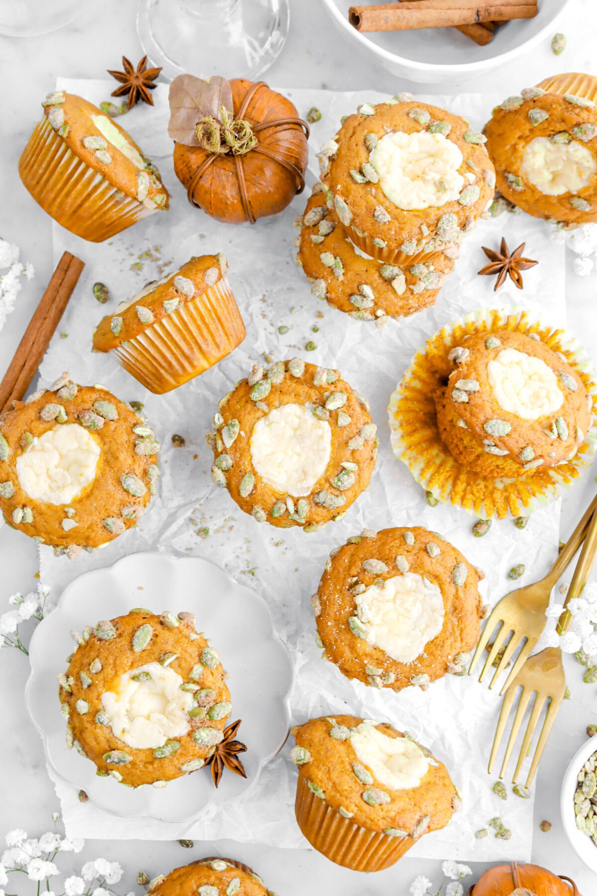 overhead image of pumpkin muffins on parchment paper with one muffin on small scalloped plate with cinnamon stick and star anise around, two gold forks, and white flowers around.