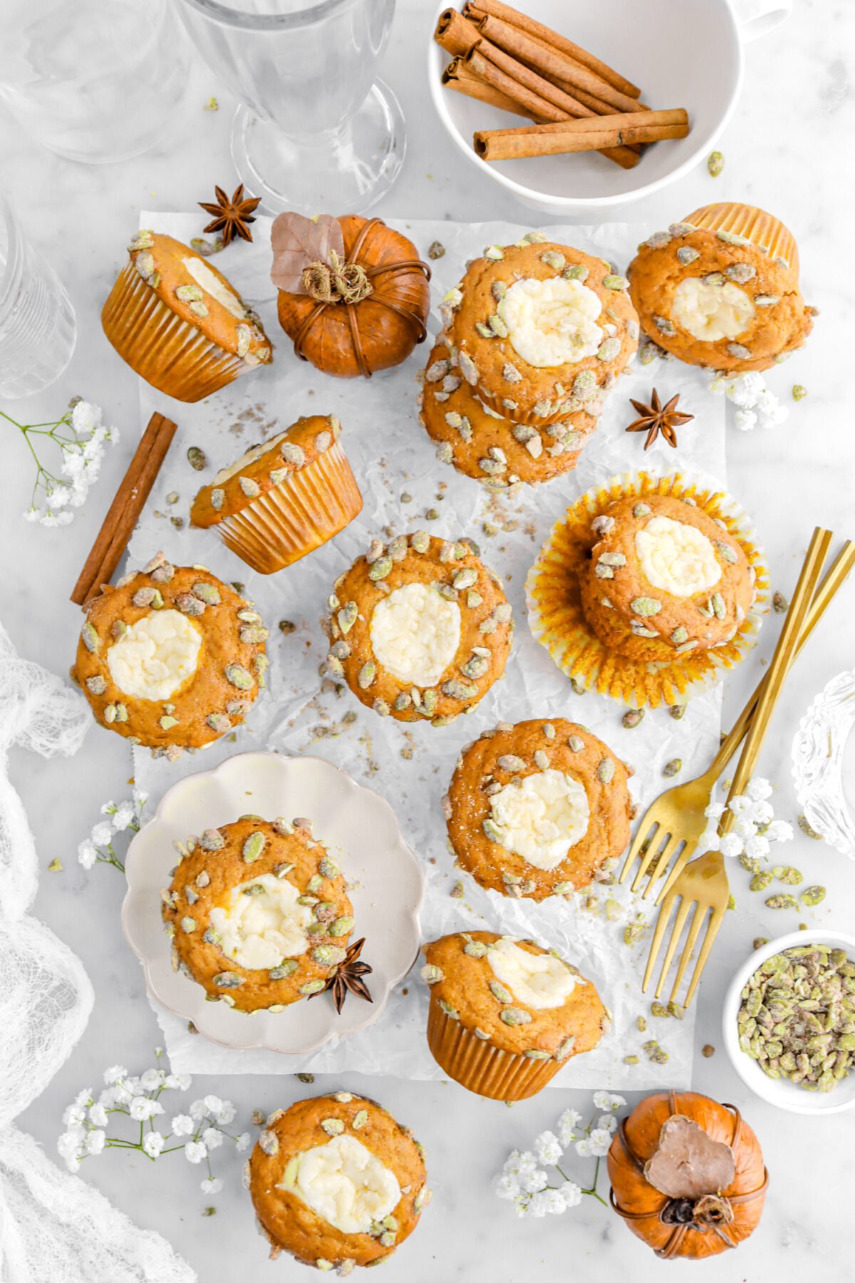 overhead image of twelve pumpkin muffins on parchment paper with one muffin on small plate with forks, faux pumpkins, white flowers, whole spices, and candied pumpkin seeds around.