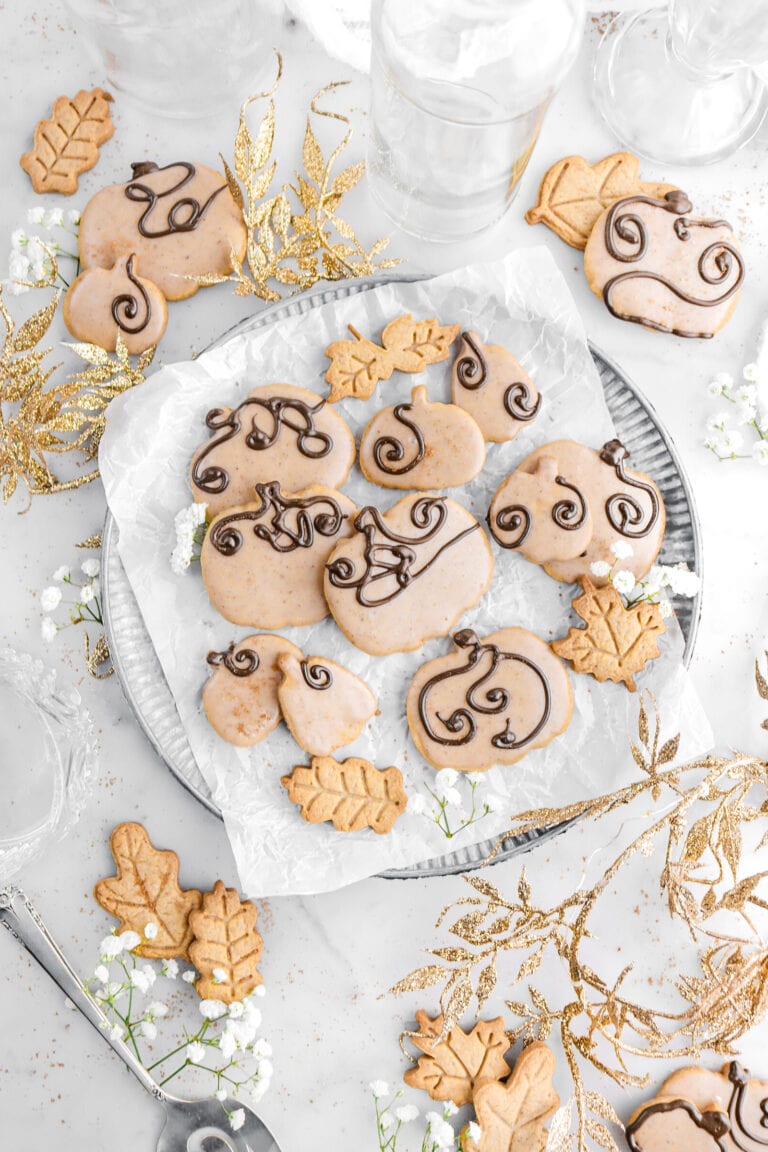 pumpkin shaped cookies on metal plate with white flowers, more cookies, and gold garland around on marble surface.