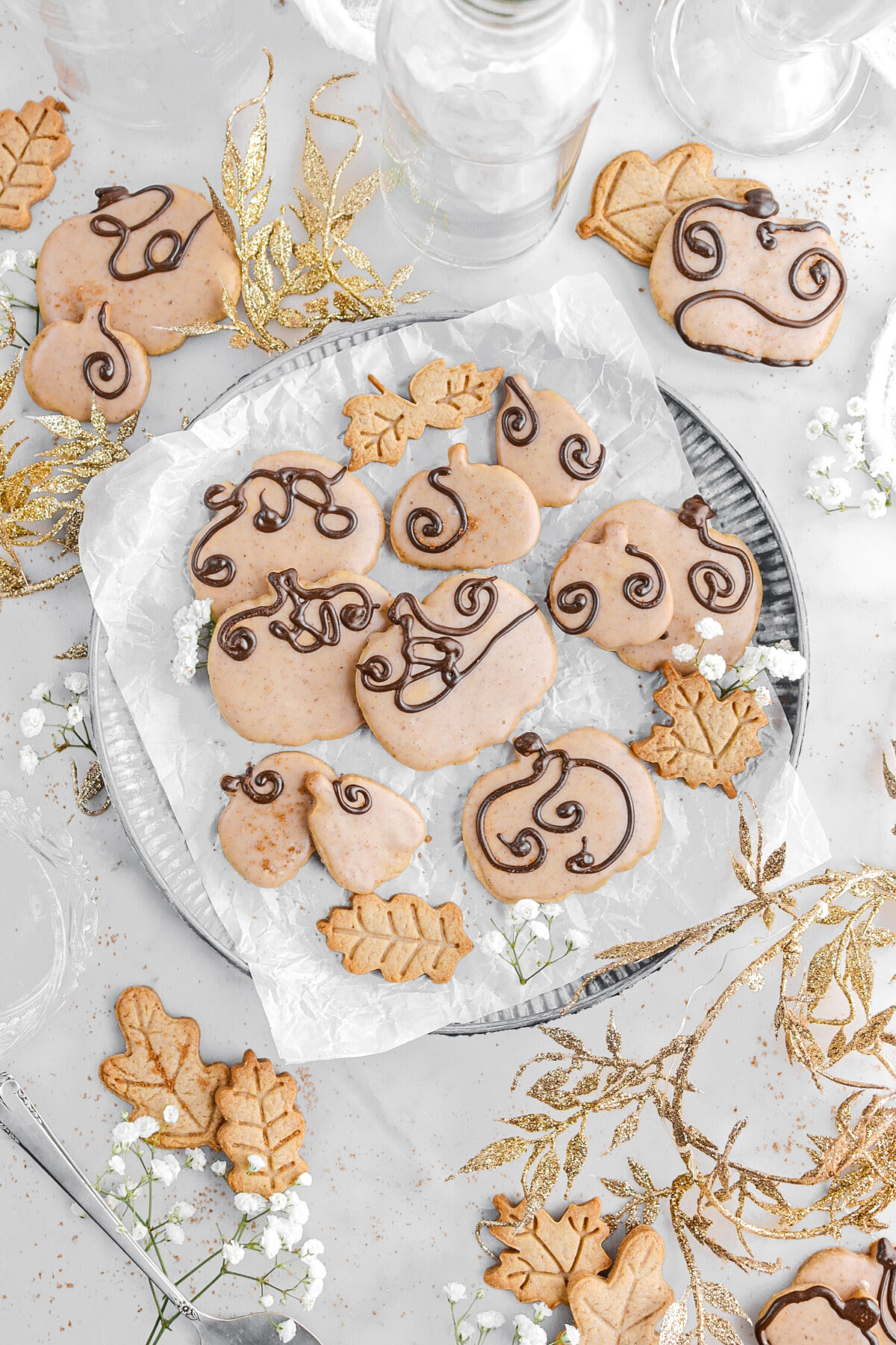 overhead image of ten pumpkin cookies on parchment paper on top of a metal plate with gold garland around and white flowers.