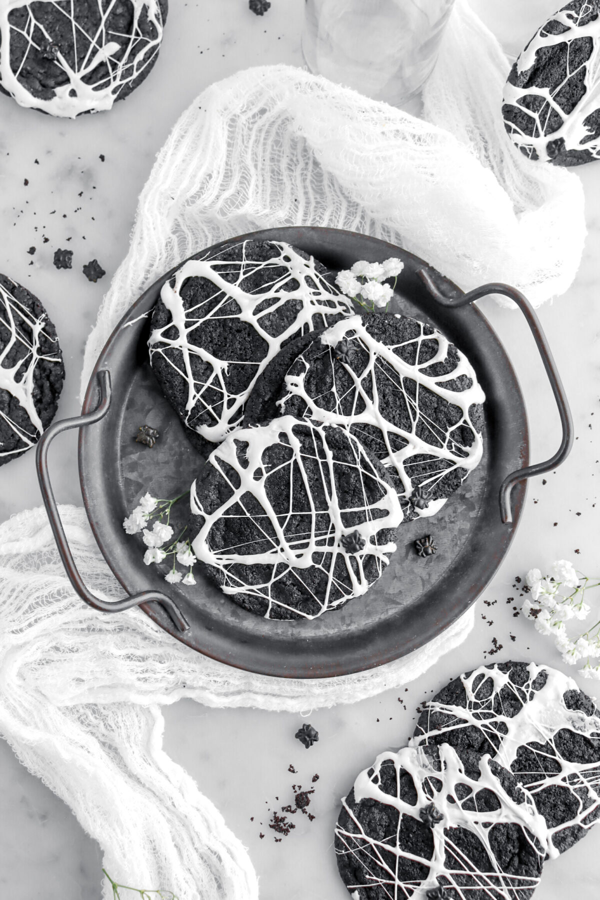 overhead image of 3 spider web cookies on metal tray on top of cheesecloth on marble surface with more cookies and white flowers around.