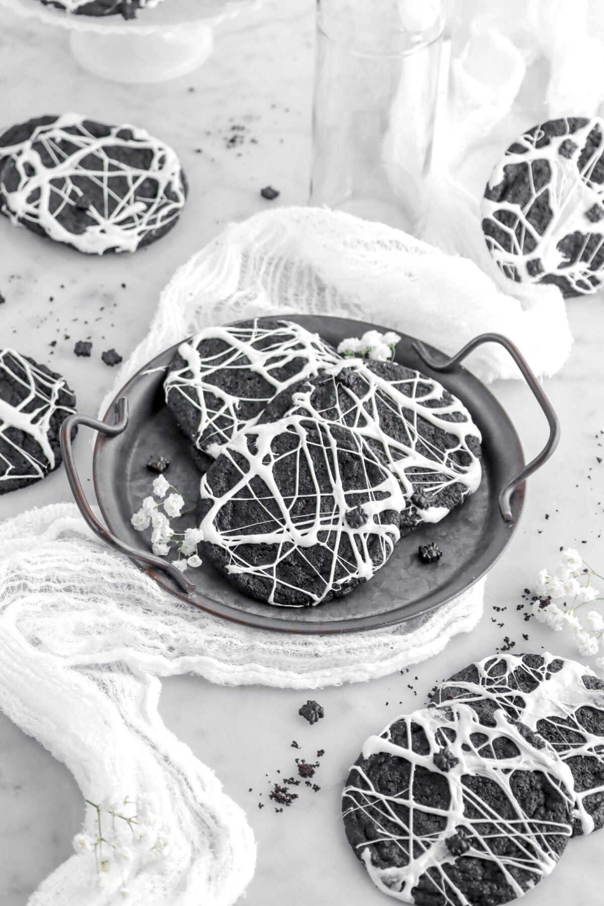 angled image of three spider web cookies on metal tray with white flowers around on marble surface.
