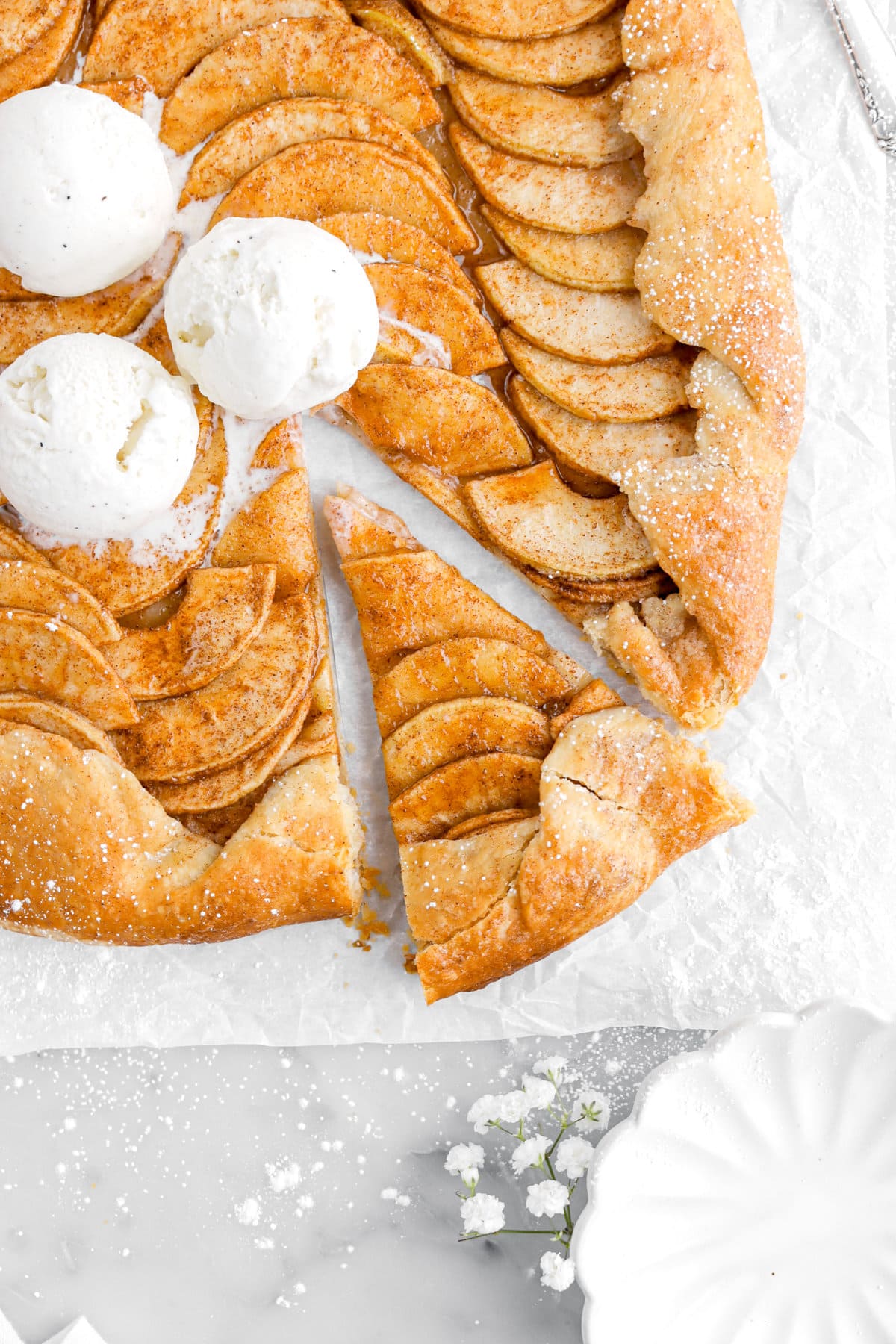 cropped overhead image of slice of apple galette on parchment paper with three scoops of ice cream on galette with bowl and flowers beside.