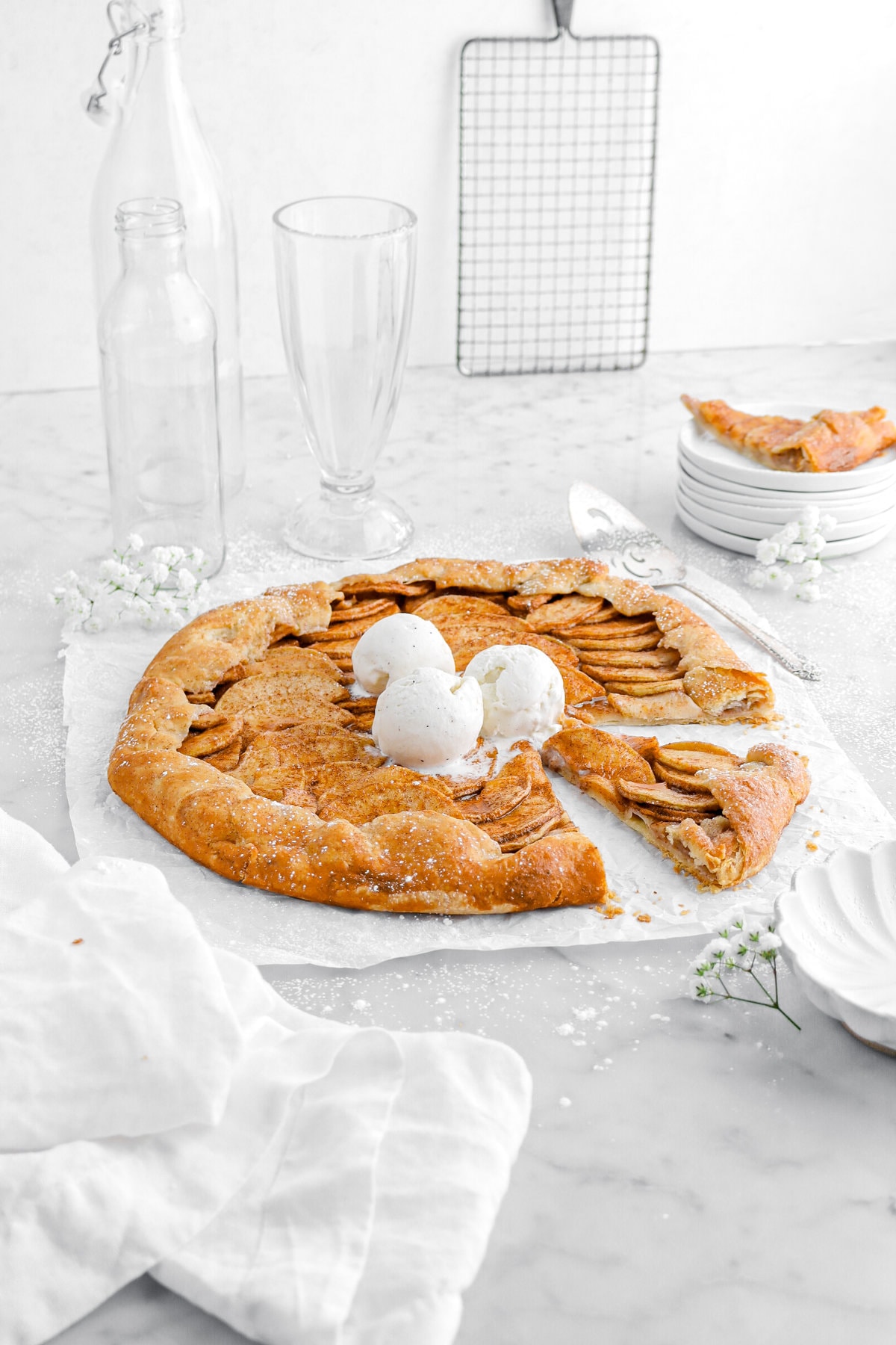 angled image of apple galette with three scoops of ice cream on parchment paper with slice cut into it and another slice behind on stack of white plates.