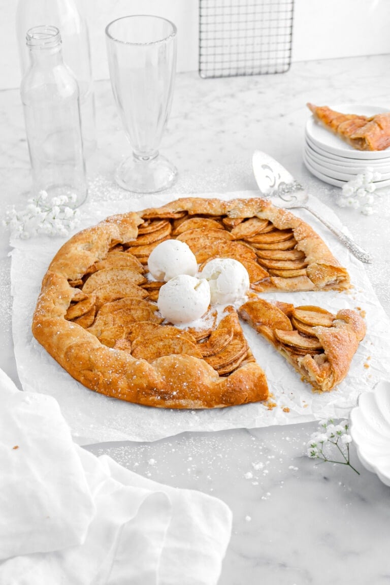 angled image of apple galette with three scoops of ice cream and slice cut into it on parchment paper with another slice of galette on stack of plates and empty glasses behind on marble surface.