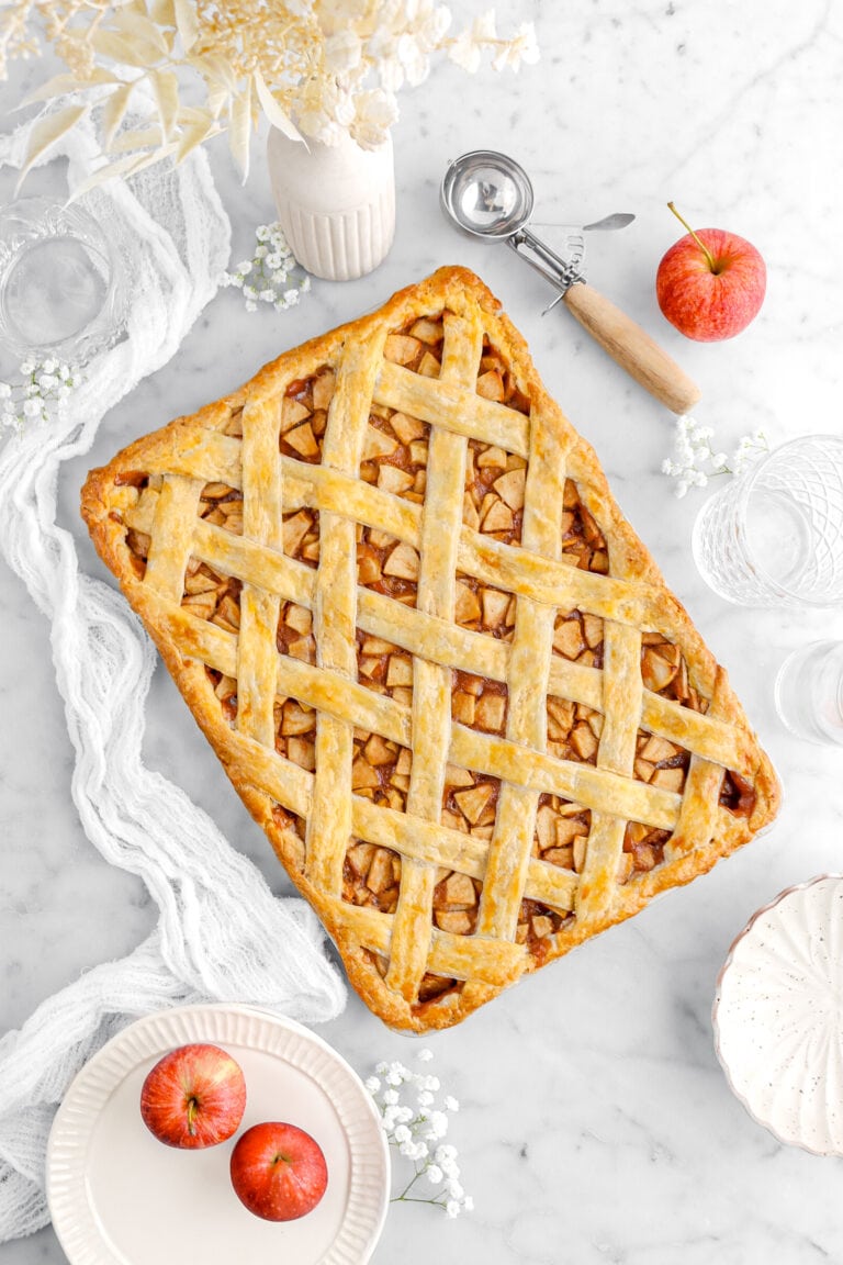 apple slab pie on marble surface with white cheesecloth beside, stack of plates, red apples, white flowers, and an ice cream scoop around on marble surface.