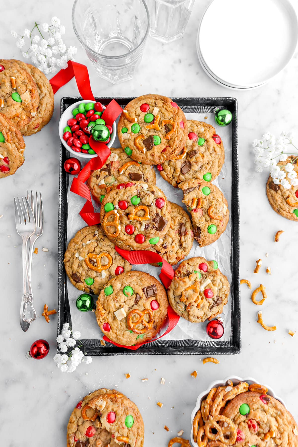 christmas monster cookies piled on lined sheet pan with red ribbon, red and green ornaments, and red and green M&M's in white bowl beside cookies with more cookies, white flowers, two forks, a plate, and a bowl of pretzels around on marble surface.