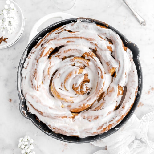 overhead image of iced cinnamon roll in skillet with white flowers, cinnamon sticks, and a white cheesecloth around.