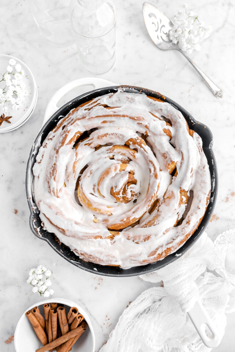 overhead image of iced cinnamon roll in skillet with white flowers, cinnamon sticks, and a white cheesecloth around.