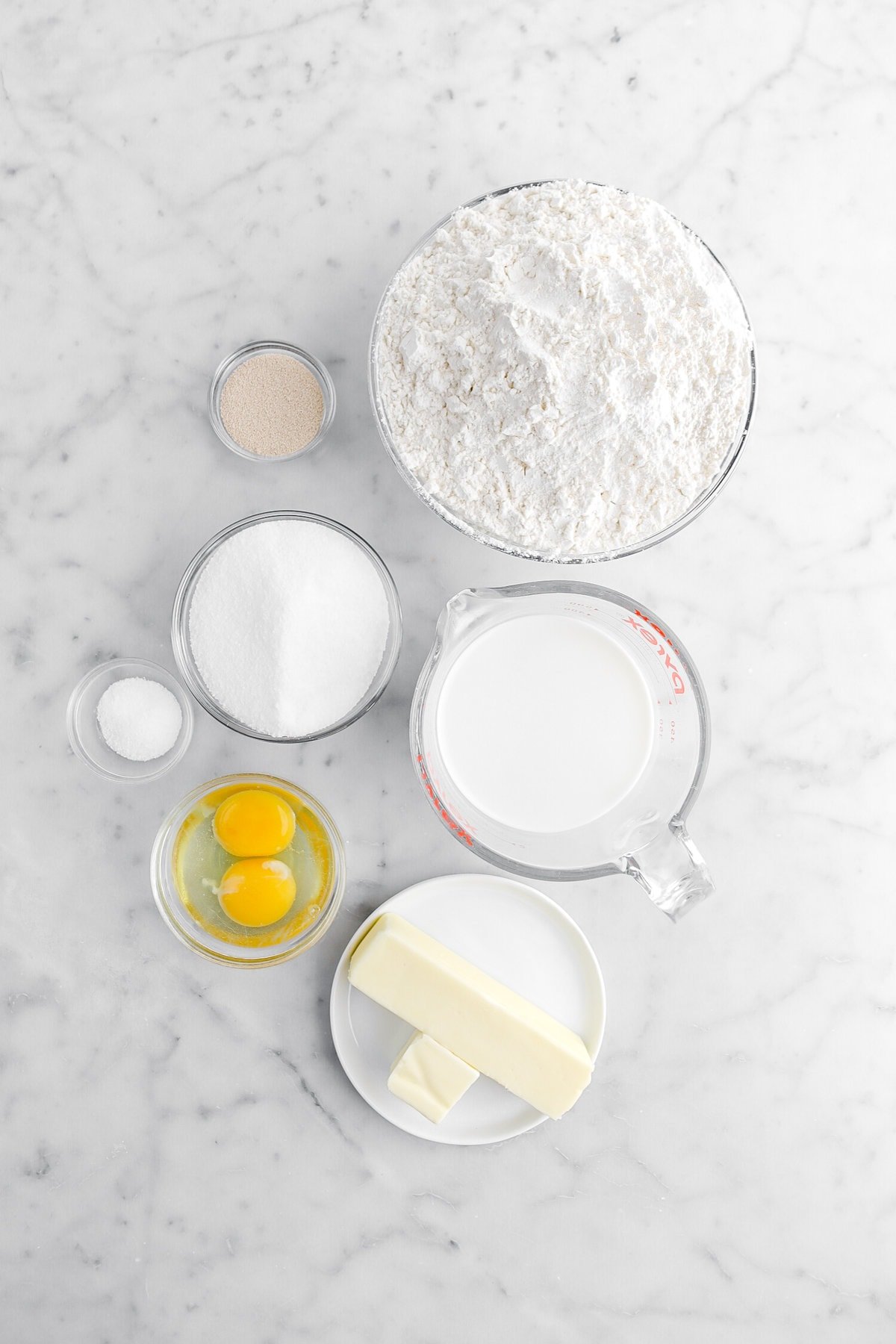 yeast, flour, sugar, salt, milk, eggs, and butter on marble surface.