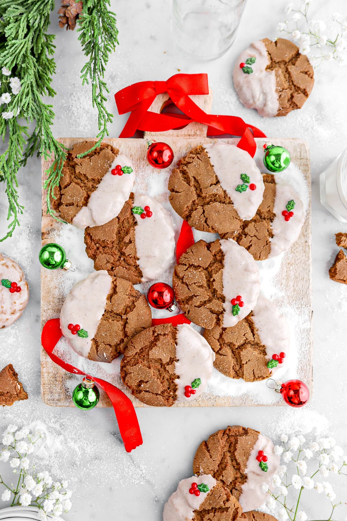 overhead image of gingerbread crinkle cookies on square wood board with mini green and red ornaments around, red ribbon, greenery, and white flowers around on marble surface.