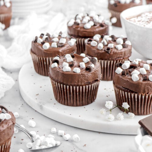 five hot chocolate cupcakes on marble serving board with mug of cocoa beside and more cupcakes around.