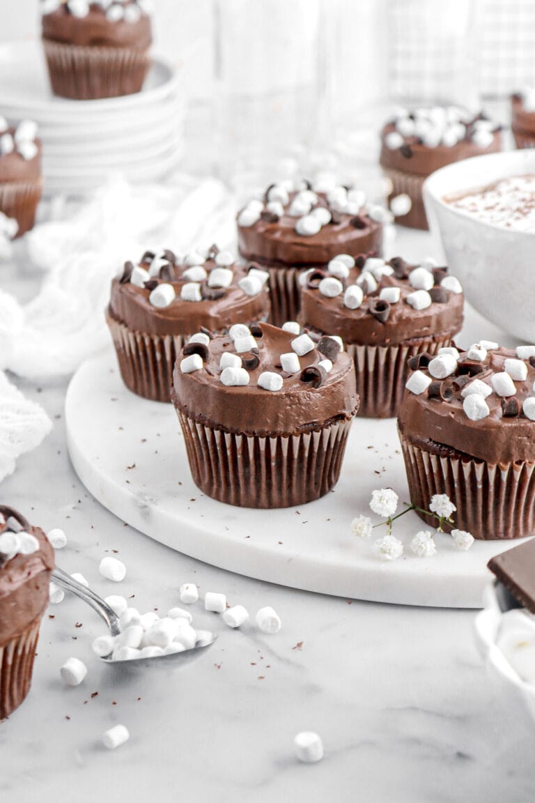 five hot chocolate cupcakes on marble serving board with mug of cocoa beside and more cupcakes around.