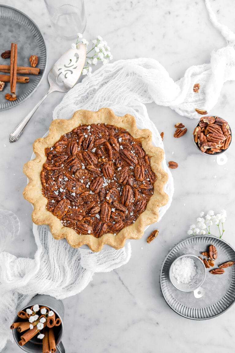pecan pie with flaked salt on top on a white cheesecloth with pecans and metal plates around on marble surface.