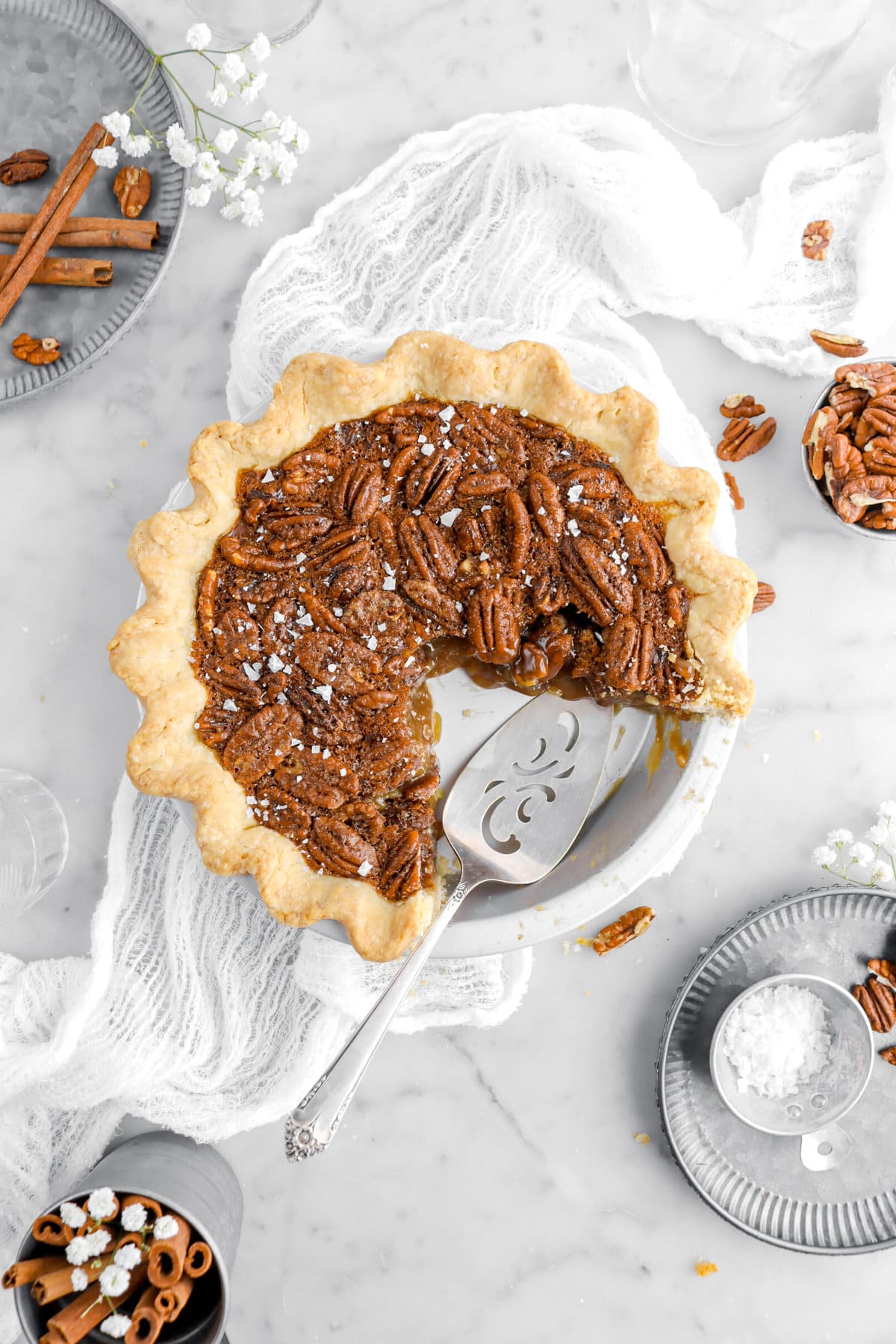 cut pecan pie in metal pi pan with cake knife placed at an angle with tip of cake knife underneath pie, metal plates, pecans, and cinnamon sticks around.