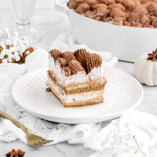 angled image of slcie of pumpkin tiramisu on white plate on top of white cheese cloth on marble surface with casserole behind and gold fork beside.