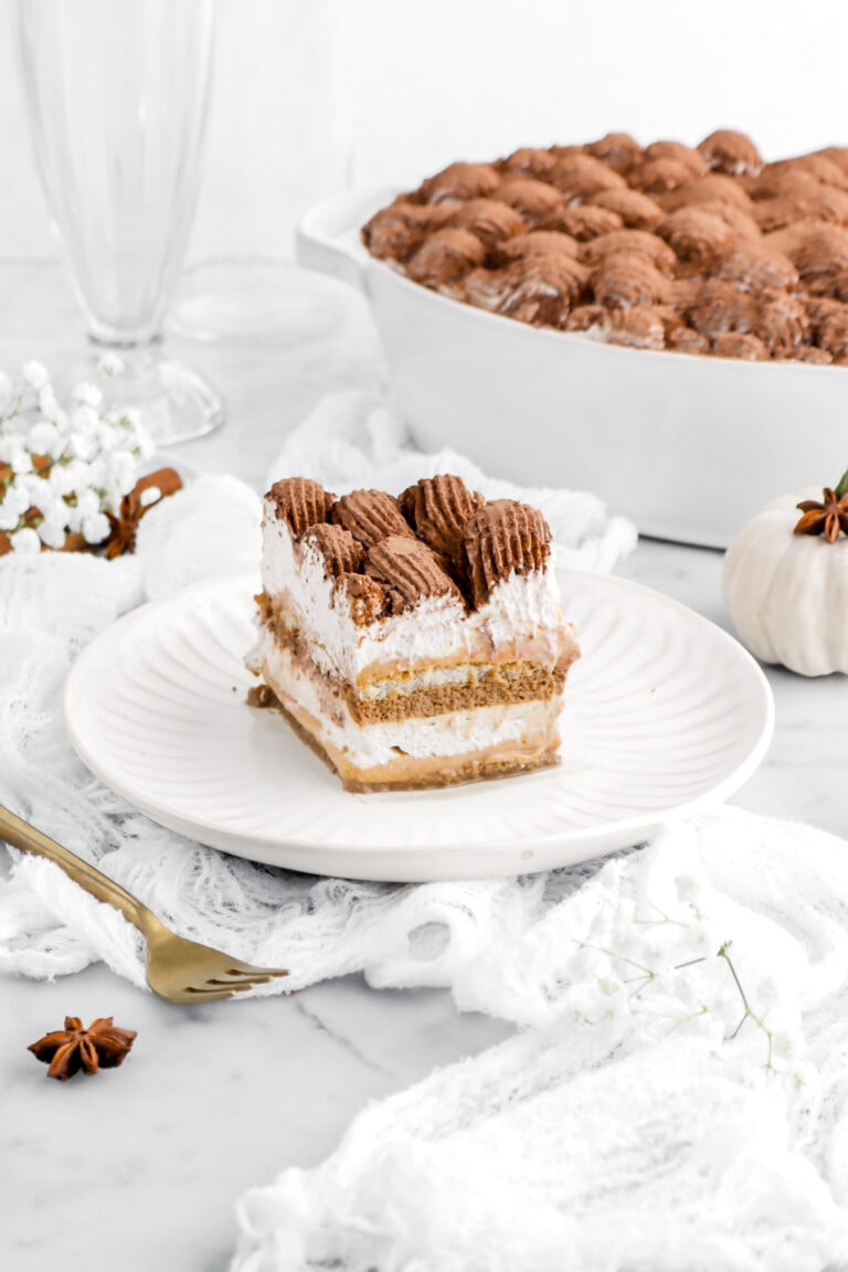 angled image of slcie of pumpkin tiramisu on white plate on top of white cheese cloth on marble surface with casserole behind and gold fork beside.