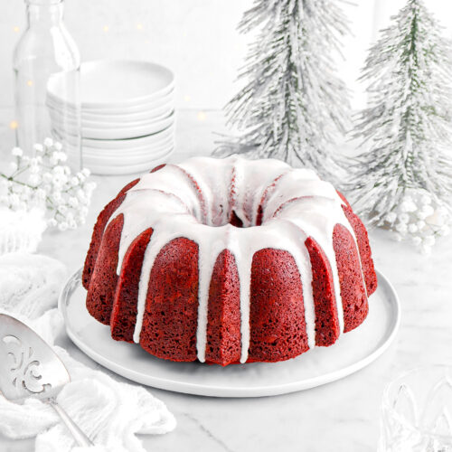 red velvet bundt cake on white plate with green christmas trees, white cheesecloth beside, and stack of white plates behind.