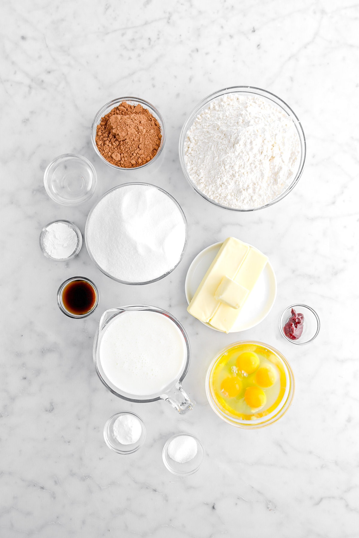 flour, cocoa powder, vinegar, sugar, baking soda, vanilla, butter, red food dye, milk, eggs, baking powder, and salt on marble surface.