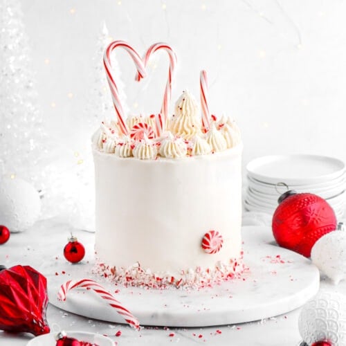 white chocolate peppermint layer cake with candy canes on top and pipped frosting with red ornaments around on marble surface.