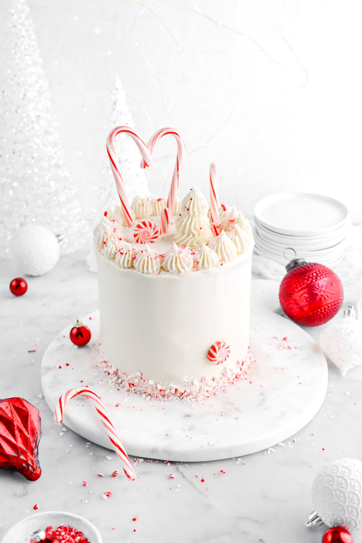 pulled back angled image of layer cake on marble surface with red and white ornaments around and crushed peppermint.