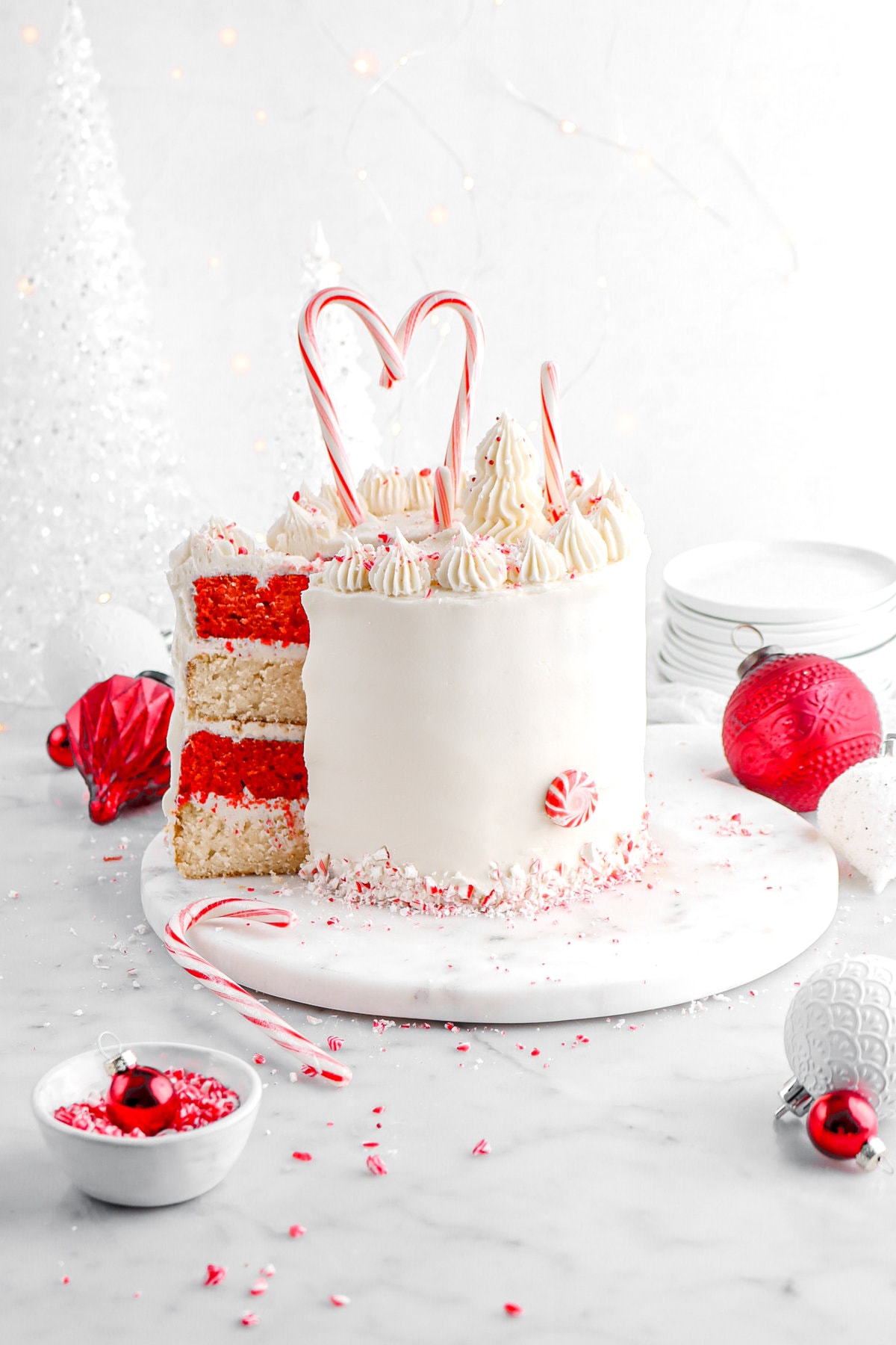 slice of white chocolate peppermint cake with red and white layers slightly pulled away from cake on marble surface with candy cane pieces and red and white ornaments around.