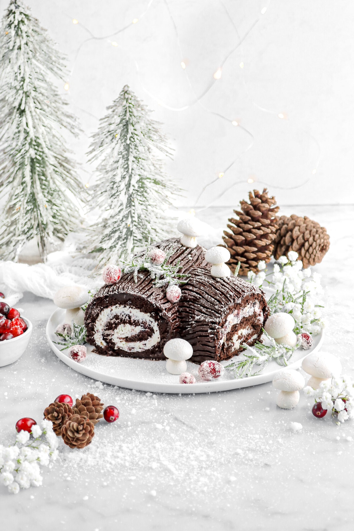 yule log on white plate with meringue mushroomed, sugared rosemary, cranberries, and white flowers around on marble surface with two mini trees behind and fairy lights.
