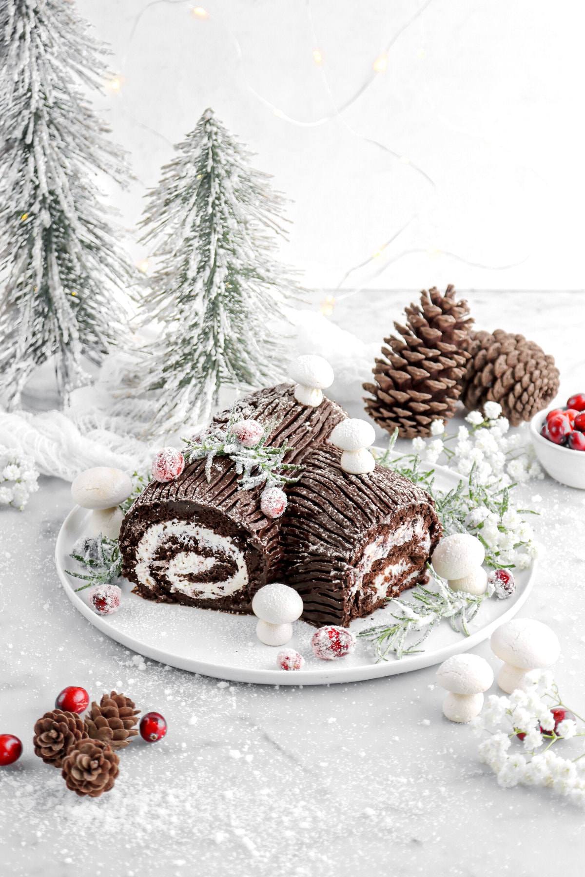 angled image of chocolate yule log on white plate with sugared cranberries and rosemary around, meringue mushrooms, white flowers, and granulated sugar around.