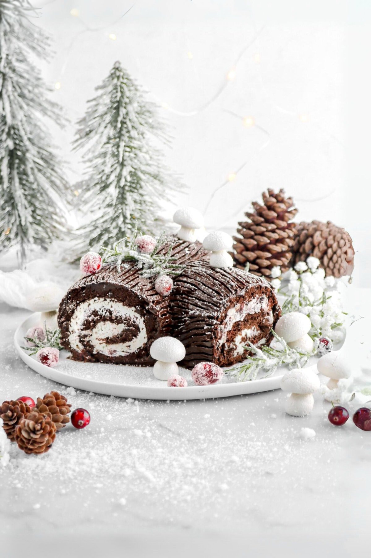 yule log on white plate with sugared rosemary and cranberries, meringue mushrooms, and pine cones around.
