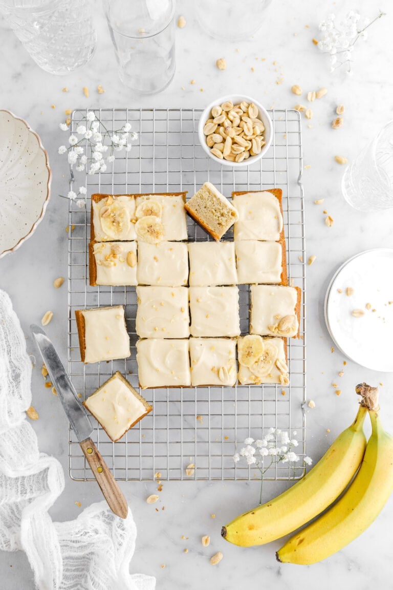overhead image of cut banana snack cake with one slice turned on it's side on wire cooling rack with white flowers, bowl of peanuts, two bananas, and a knife around on marble surface.