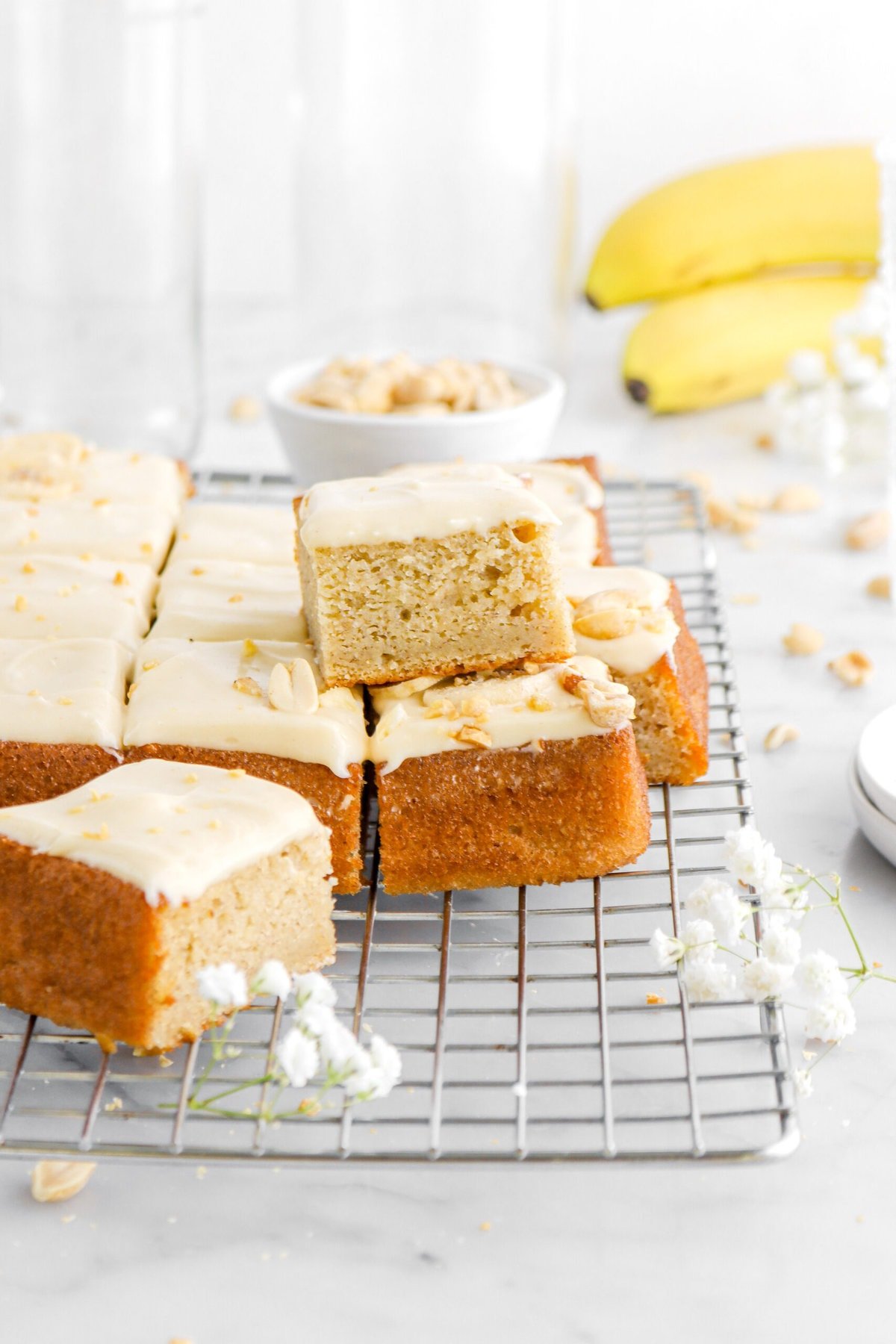 slice of banana cake stacked on top of cut cake on a wire cooling rack with white flowers in front, a bowl of peanuts and bananas behind.