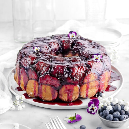 lemon blueberry monkey bread on white plate with flowers, blueberries, forks, and white plates around on marble surface.