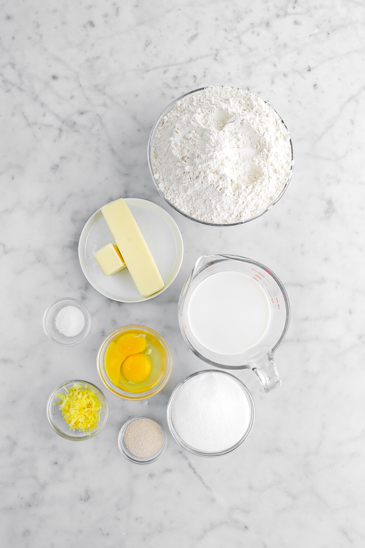 flour, butter, milk, salt, eggs, lemon zest, yeast, and sugar on marble surface.