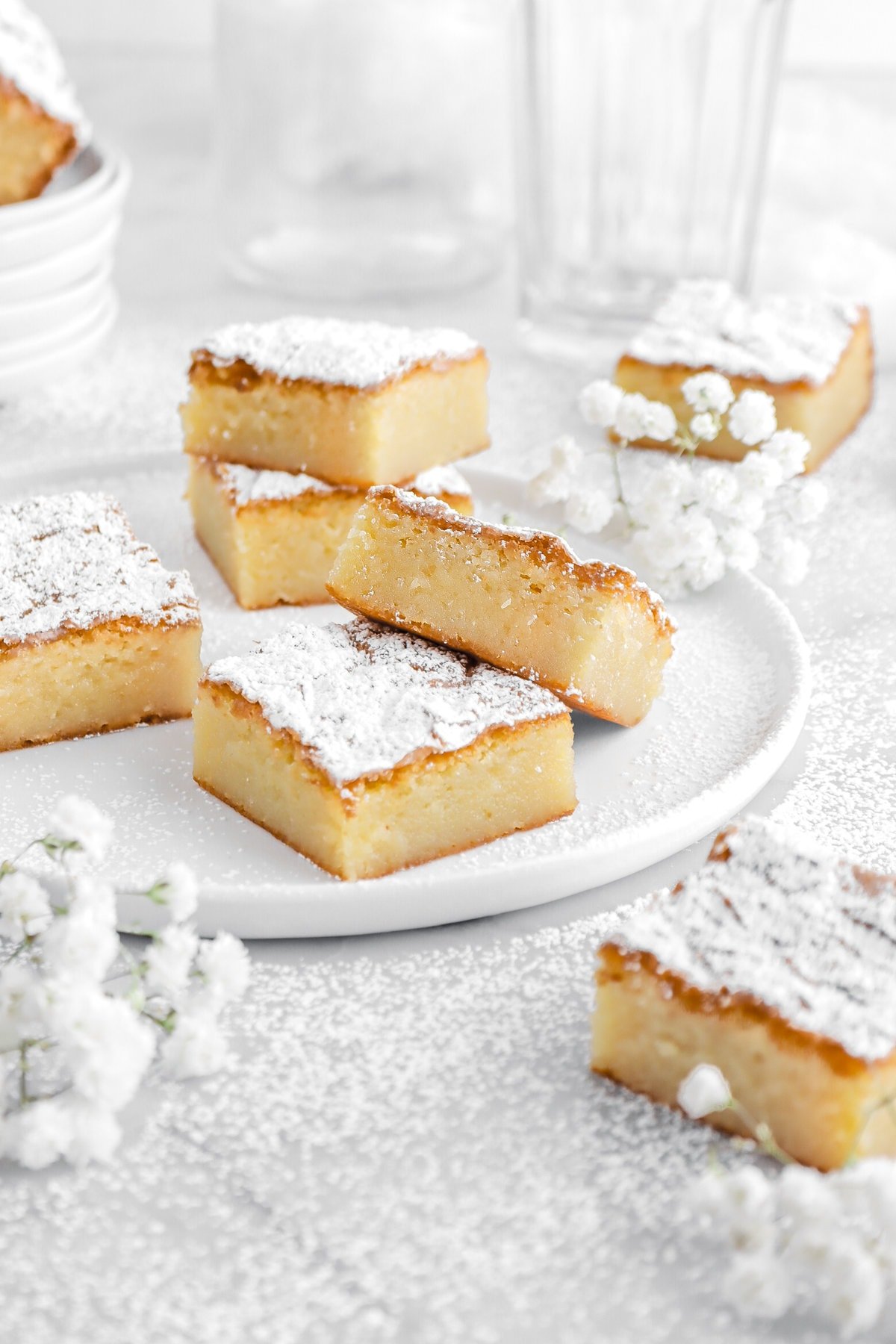 two white chocolate brownies propped against each other on white plate with more rownies around and white flowers.