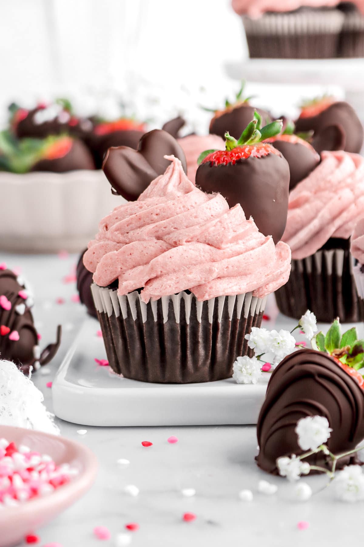 chocolate cupcake with strawberry frosting on white tray with more cupcakes behind and bowl of chocolate dipped strawberries.