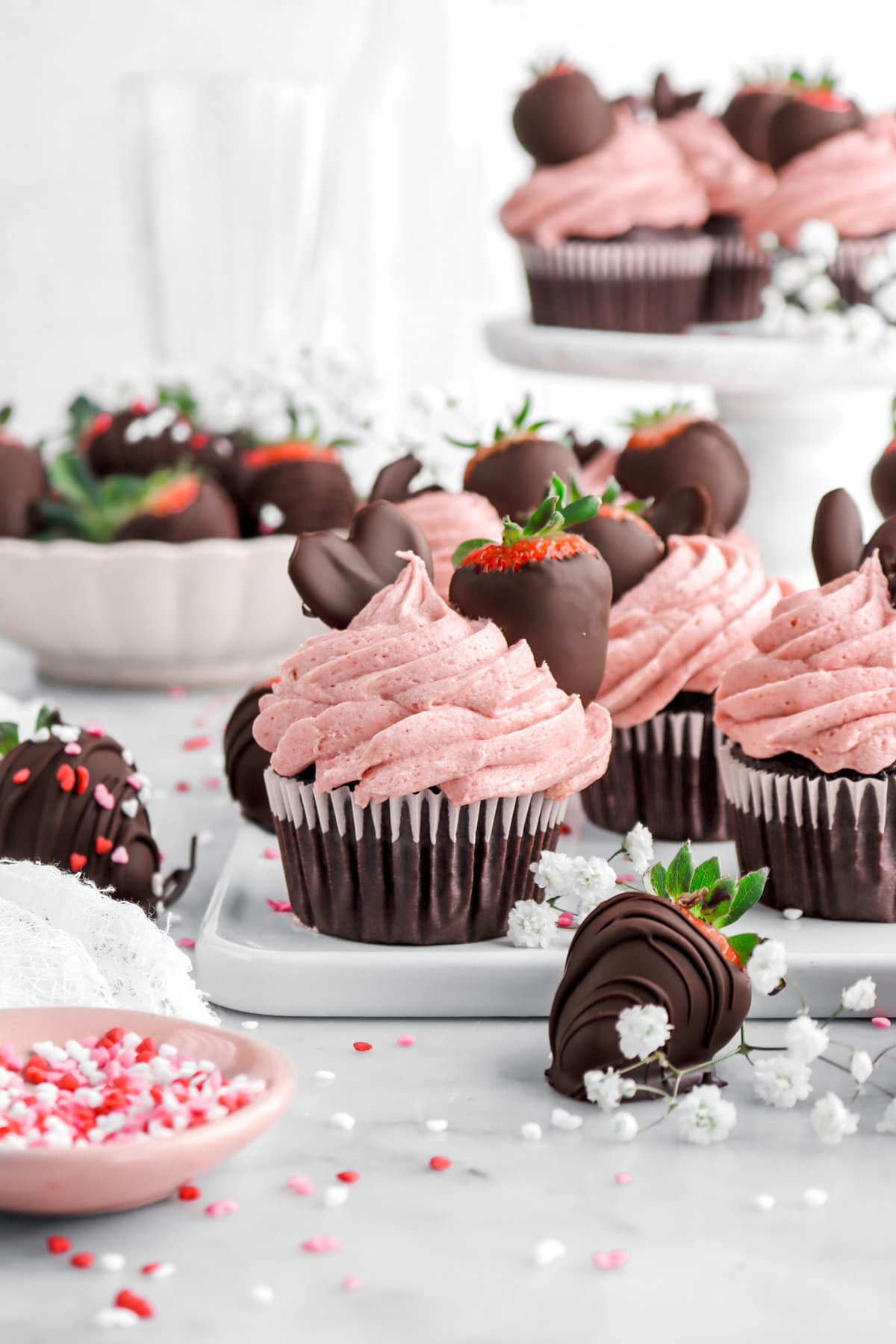 front image of chocolate covered strawberry cupcake with more cupcakes behind, as well as chocolate covered strawberries and mini heart sprinkles around.